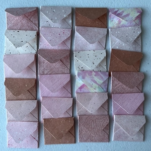 Puede incluir: Una colección de veinte sobres cuadrados pequeños dispuestos en una cuadrícula de cuatro por cinco. Los sobres presentan diversas texturas y patrones en tonos rosa, marrón, crema y blanco, incluyendo diseños moteados, marmolados y teñidos.