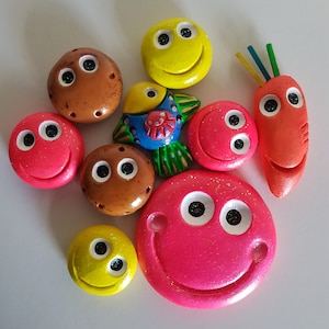 May include: A collection of nine colorful, hand-painted, ceramic smiley face ornaments. The ornaments are in various shapes and sizes, including a fish, a carrot, and a round shape. The ornaments are all smiling and have different colored eyes.