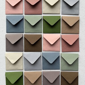 Puede incluir: Una colección de veinte sobres de papel cuadrados dispuestos en una cuadrícula de cuatro por cinco. Cada sobre presenta una solapa triangular y viene en un color mate distinto, incluyendo tonos de rosa, azul, verde, beige, gris y marrón.