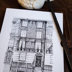 Puede incluir: Dibujo a tinta en blanco y negro de una fachada de edificio con exterior de ladrillo, ventanas y una valla de hierro forjado. También se ven una varita decorativa y una taza con forma de búho.