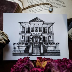 Peut inclure: Illustration architecturale en noir et blanc d'un grand bâtiment avec des colonnes, des balcons et une clôture en fer forgé. L'image est sur une carte blanche, avec des pétales de rose séchés au premier plan et un collier et un livre en arrière-plan. Le texte "britsbazaar" est en bas.