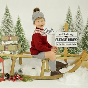Può includere: Un bambino piccolo con un maglione rosso e un cappello di lana grigio è seduto su una slitta di legno. È circondato da un paesaggio invernale innevato con pini e un cartello che dice "Old Fashioned Sledge Rides Hot Chocolate & Jingle Bells".