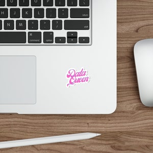 May include: A white sticker with the text "Data Queen" in a retro, cursive font, shaded in pink and purple. The sticker is placed on a laptop, with a wooden desk and a computer mouse in the background.