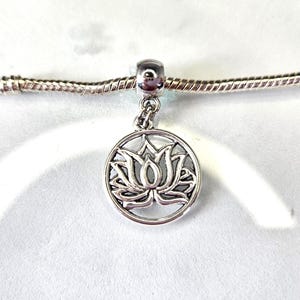Peut inclure: Un charm argenté avec un motif de fleur de lotus. Le charm est rond avec un motif ajouré et est suspendu à une perle argentée. Le charm est attaché à un bracelet argenté.