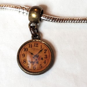 May include: A vintage-style charm featuring a clock face with black hands and numerals on a tan background. The charm is round with a bronze-colored frame and is suspended from a silver-colored chain. The clock shows the time as approximately 11:12.