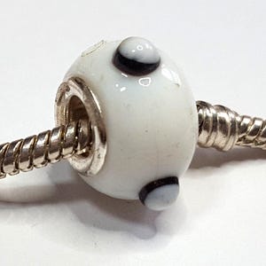 May include: A white glass bead with three black and white accents. The bead is on a silver bracelet. The bead has a smooth, glossy finish and is cylindrical in shape. The bead is a charm.
