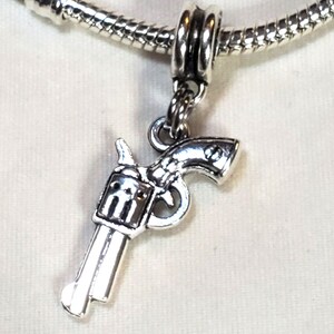 May include: A silver-colored charm in the shape of a revolver pistol. The charm is suspended from a silver-colored snake chain. The pistol charm has detailed features, including the trigger, hammer, and barrel.