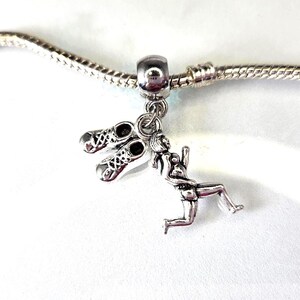 Könnte beinhalten: Silberner Charm mit einem Paar Laufschuhen und einer Läuferfigur. Der Charm ist an einer silbernen Kette befestigt. Die Laufschuhe haben detaillierte Schnürsenkel. Der Läufer befindet sich in einer Laufposition.