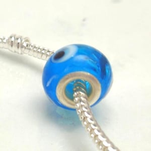 May include: A bright blue glass bead with a circular evil eye pattern featuring white and black details, threaded onto a silver-toned snake chain bracelet. The bead has a metallic core and a smooth, rounded surface.