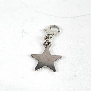 May include: A silver-colored star charm with a lobster clasp. The five-pointed star is made of a shiny metal and is suspended from a small clasp. The charm is designed to be attached to a bracelet or necklace.
