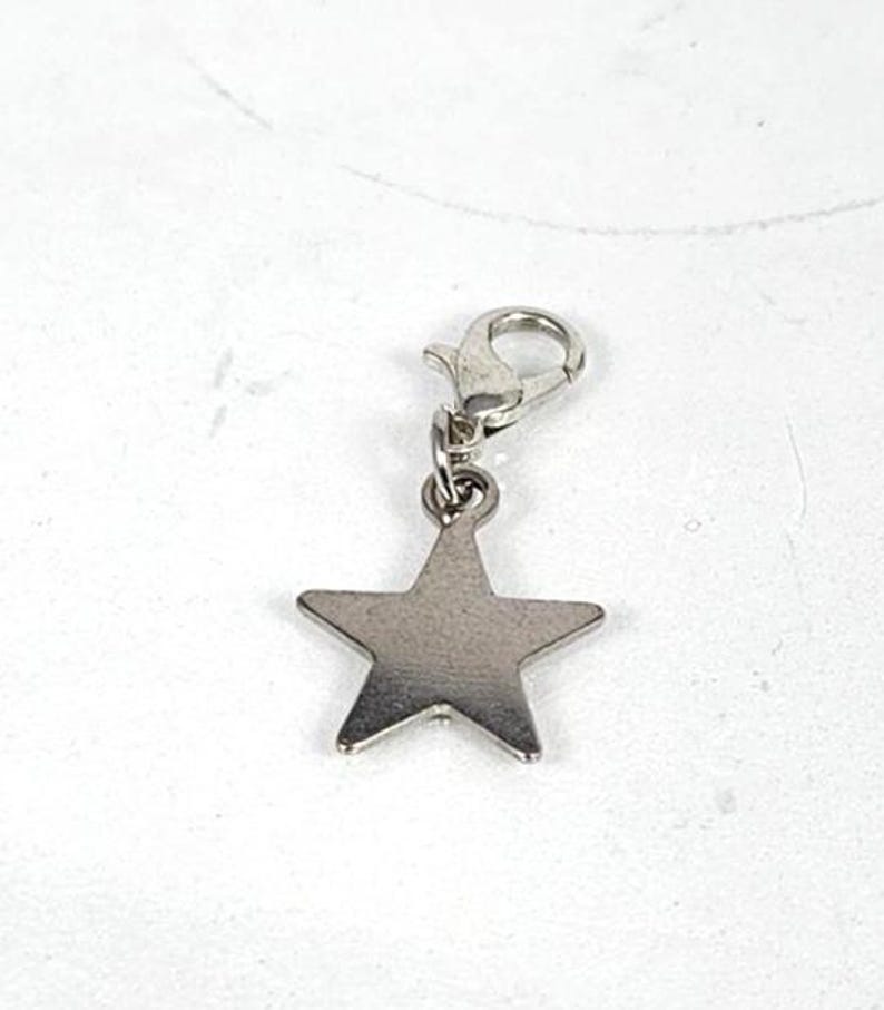 May include: A silver-colored star charm with a lobster clasp. The five-pointed star is made of a shiny metal and is suspended from a small clasp. The charm is designed to be attached to a bracelet or necklace.