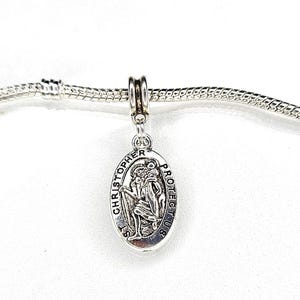 Könnte beinhalten: Silbernes Charm-Armband mit einem silbernen Anhänger, der eine Darstellung des Heiligen Christophorus mit dem Text "St. Christopher Protect Us" zeigt.