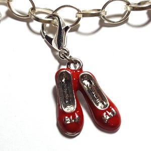 Puede incluir: Una pulsera de plata con un par de zapatos de esmalte rojo. Los zapatos son de color rojo brillante con detalles plateados y están unidos a un cierre plateado. El dije es una representación en miniatura de zapatos rojos.