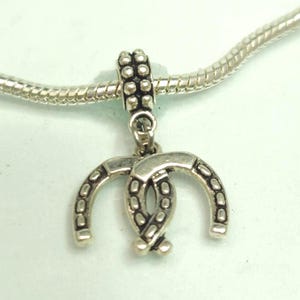 May include: Silver-toned charm featuring two horseshoes intertwined, suspended from a beaded bail. The charm is attached to a silver snake chain, creating a jewelry piece. The horseshoe charm is a symbol of good luck.