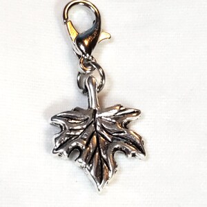 May include: Silver-toned charm featuring a detailed maple leaf design. The charm is attached to a lobster clasp, allowing it to be easily added to a necklace, bracelet, or keychain. The leaf has intricate veining.