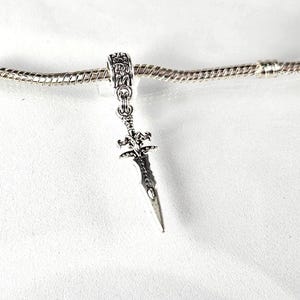 May include: Silver charm in the shape of a sword with intricate details. The charm is attached to a silver chain.
