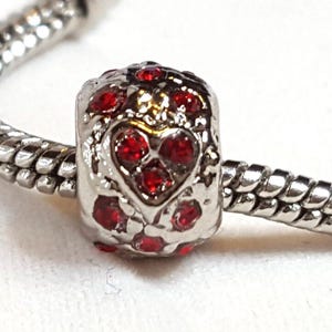 May include: A silver charm bead with a heart design, embellished with small red gemstones. The charm is on a silver snake chain bracelet, perfect for adding to a charm bracelet. The bead has a textured surface and a heart-shaped design.