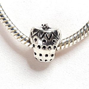 May include: A silver charm with a detailed design, resembling a small decorative object. The charm features a zig-zag pattern and small circular cutouts. It is strung on a silver coiled chain, suitable for bracelets or necklaces.