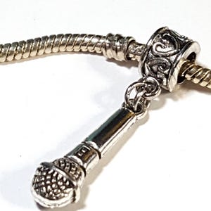 May include: A silver-toned charm featuring a miniature microphone pendant. The charm is attached to a silver snake chain. The microphone has a detailed, textured head and a sleek, cylindrical body. The charm is designed for jewelry making.