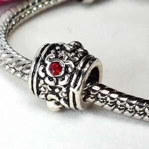 May include: A silver charm bead with an ornate floral design and a small red gemstone. The bead is on a silver snake chain, creating a bracelet or necklace component. The charm is detailed and decorative.