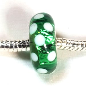 May include: A green glass bead with white polka dots. The bead is cylindrical and sits on a silver-colored metal bracelet. The bead has a shiny, reflective surface and is a decorative element.