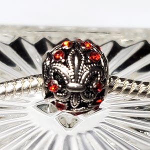 Può includere: Un ciondolo sferico in argento con un intricato design in rilievo a forma di giglio, impreziosito da piccoli cristalli rotondi di un rosso vibrante. Il ciondolo è infilato su un bracciale a catena snake in argento con finitura testurizzata.