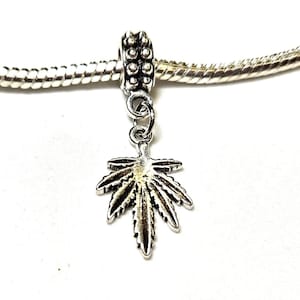 May include: A silver-colored charm featuring a detailed cannabis leaf design. The charm is suspended from a beaded silver-tone bead and a silver-tone chain. The charm is designed to be added to a bracelet or necklace.