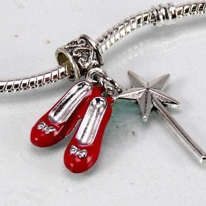 Op de afbeelding: Een zilverkleurige bedelarmband met twee bungelende bedels: een paar felrode ballerina's met kleine zilveren strikjes en een zilveren ster-vormige toverstaf. De bedels zijn bevestigd aan een flexibele, getextureerde zilveren slangenketting.