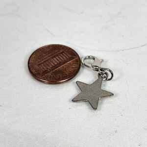 May include: A silver-colored star charm with a lobster clasp next to a U.S. penny. The star charm is a five-pointed shape. The penny is bronze and shows the profile of Abraham Lincoln.