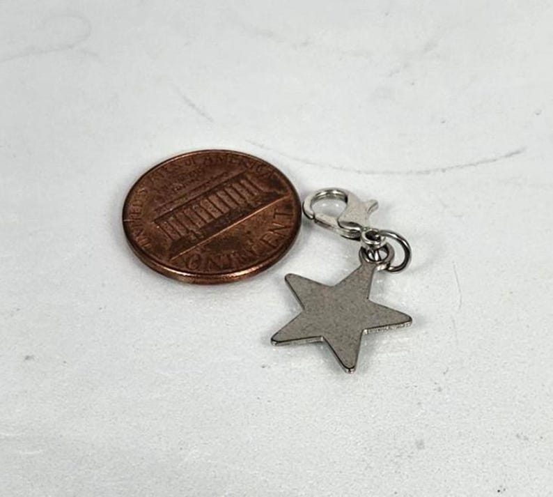 May include: A silver-colored star charm with a lobster clasp next to a U.S. penny. The star charm is a five-pointed shape. The penny is bronze and shows the profile of Abraham Lincoln.