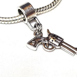 May include: Silver-toned charm in the shape of a revolver, suspended from a silver chain. The charm features detailed design elements, including the barrel, trigger, and hammer. The jewelry piece is set against a white background.