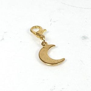 Könnte beinhalten: Ein goldfarbener Anhänger mit einem Halbmond-Anhänger. Der Anhänger hat einen Karabinerverschluss zur einfachen Befestigung an einem Armband oder einer Halskette. Der Mond-Anhänger ist ein schlichtes, elegantes Design.