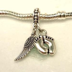 Könnte beinhalten: Ein silberner Charm-Anhänger mit einem detaillierten Flügel und einem Paar Babyfüßen. Der Anhänger hängt an einem Spacer mit Perlen und ist für ein Armband oder eine Halskette gedacht. Der Anhänger ist aus glänzendem Metall gefertigt.