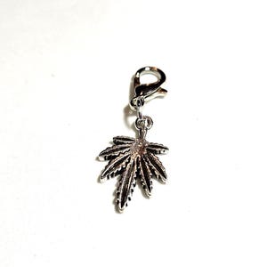 Peut inclure: Un charm argenté représentant une feuille de cannabis détaillée. Le charm est attaché à un fermoir mousqueton, permettant de l'ajouter facilement à un collier, un bracelet ou d'autres bijoux. La feuille présente des nervures complexes et une surface texturée.