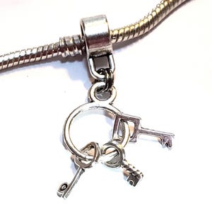 May include: A silver-toned charm featuring a circular ring with three small dangling keys attached, suspended from a cylindrical bail on a matching silver-toned snake chain necklace. The metal components have a polished, metallic finish.