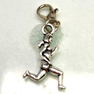 Könnte beinhalten: Ein silberfarbener Metallanhänger in Form einer laufenden Person mit glatter, reflektierender Oberfläche und einem kleinen Karabinerverschluss an der oberen Öse, der zur Befestigung an Schmuckstücken wie Armbändern oder Halsketten dient.