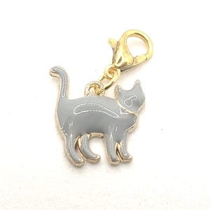 May include: A gray metal charm of a cat with a gold-colored clasp. The charm is perfect for adding a touch of whimsy to a bag, keychain, or jewelry.