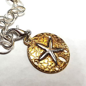 May include: A gold-coloured pendant with a silver starfish design, attached to a silver chain. The pendant has a textured surface and is round with a starfish in the centre. The chain has a lobster clasp.