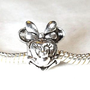 May include: A silver-toned metal charm shaped like the head of a cartoon mouse with a large bow, attached to a flexible snake chain bracelet. The charm features engraved details for the eyes, eyelashes, nose, and mouth, with a polished finish.