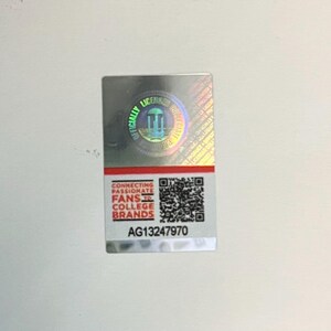 May include: A rectangular, silver sticker with a holographic sheen. The top portion features a circular seal with the words "OFFICIALLY LICENSED" and a graphic. The bottom section has text that reads "CONNECTING PASSIONATE FANS TO COLLEGE BRANDS" and a QR code. The sticker's serial number is AG13247970.