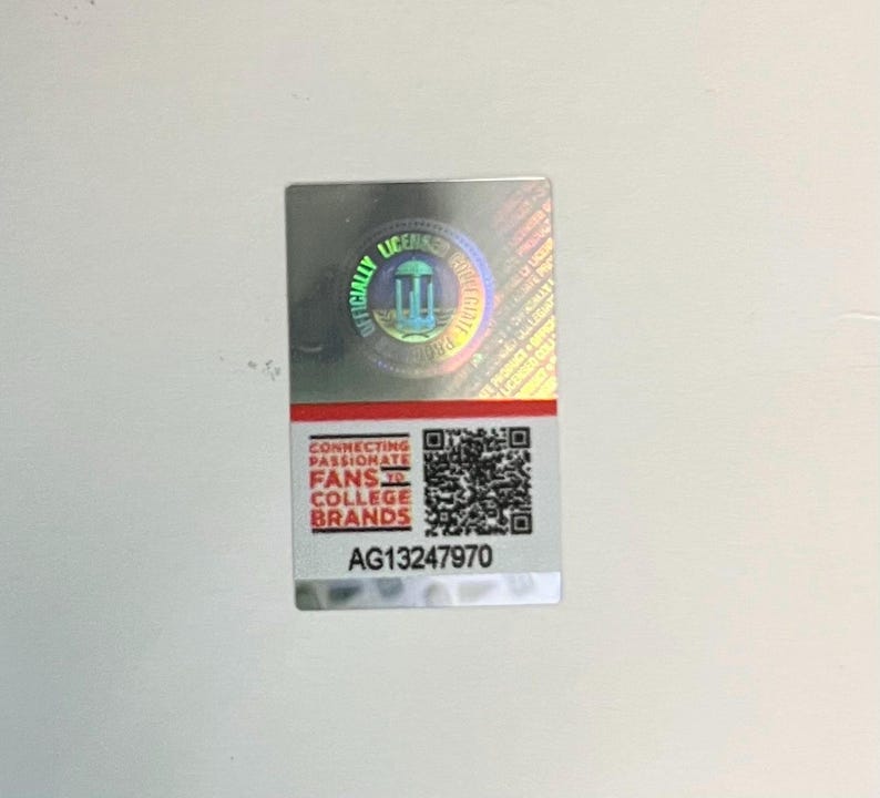 May include: A rectangular, silver sticker with a holographic sheen. The top portion features a circular seal with the words "OFFICIALLY LICENSED" and a graphic. The bottom section has text that reads "CONNECTING PASSIONATE FANS TO COLLEGE BRANDS" and a QR code. The sticker's serial number is AG13247970.