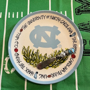 May include: A ceramic plate with a light blue rim and a light blue center featuring the University of North Carolina logo. The plate has a stainless steel cheese knife. The plate has the text "Hark the Sound" and "Carolina Fever UNC".