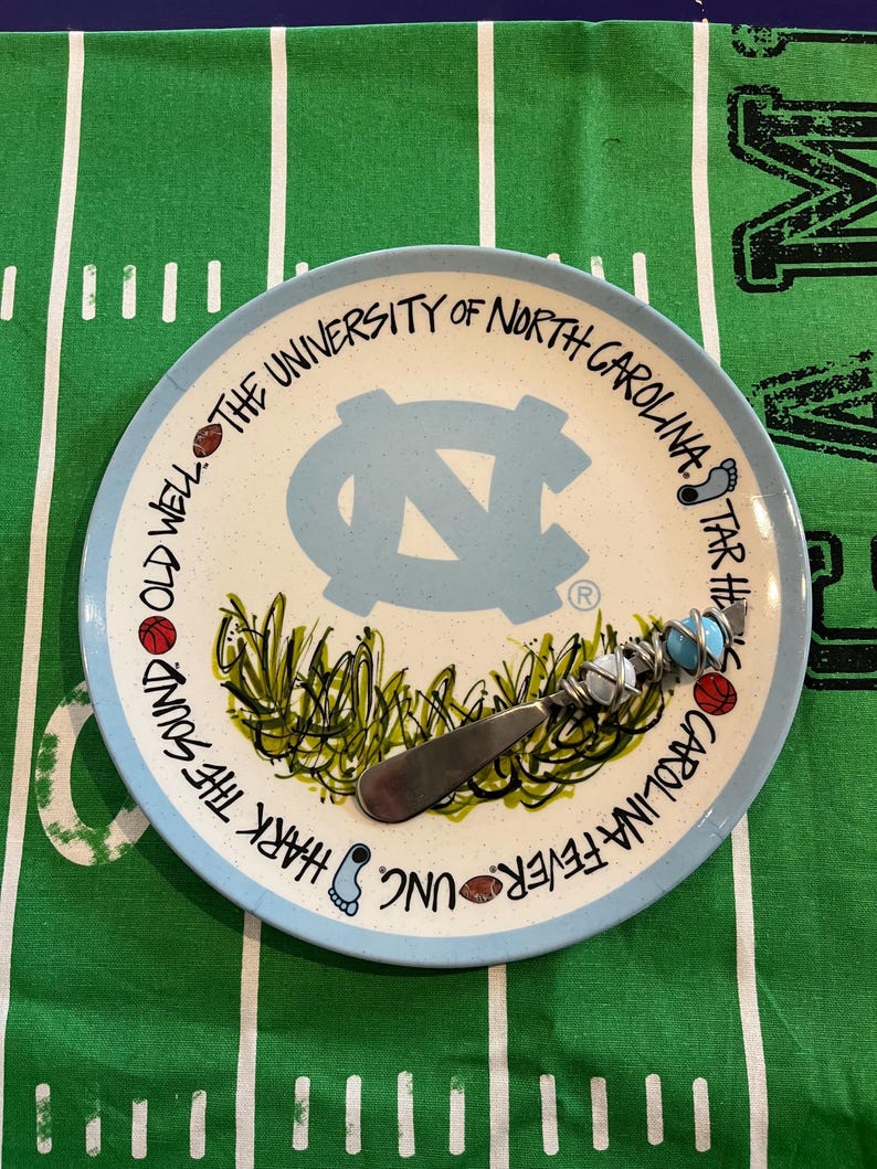 May include: A ceramic plate with a light blue rim and a light blue center featuring the University of North Carolina logo. The plate has a stainless steel cheese knife. The plate has the text "Hark the Sound" and "Carolina Fever UNC".