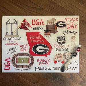 May include: A rectangular glass cutting board featuring a collage of University of Georgia Bulldogs imagery. The design includes the UGA logo, a bulldog, the state of Georgia, and various phrases in red and black.