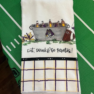 May include: White hand towel with a football theme. The towel features a drawing of a metal bucket filled with bottles, a football, and the text "Eat, Drink & Go Pirates!" The bottom of the towel has a grid pattern in purple and gold.
