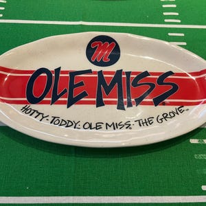 May include: Oval-shaped ceramic serving platter with a cream-colored base and red and navy blue accents. The platter features the text "OLE MISS" in navy blue, with the Ole Miss logo and the phrases "HOTTY-TODDY: OLE MISS: THE GROVE."