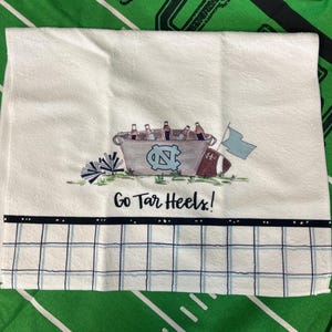 May include: White hand towel with a decorative design featuring a bucket of bottles, a football, and a flag. The towel has the text "Go Tar Heels!" and a blue and white checkered pattern.
