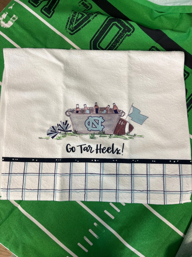 May include: White hand towel with a decorative design featuring a bucket of bottles, a football, and a flag. The towel has the text "Go Tar Heels!" and a blue and white checkered pattern.