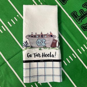 May include: White hand towel with a football field design. The towel features a bucket of beer bottles, the UNC logo, and the text "Go Tar Heels!". A black and blue checkered pattern is at the bottom.