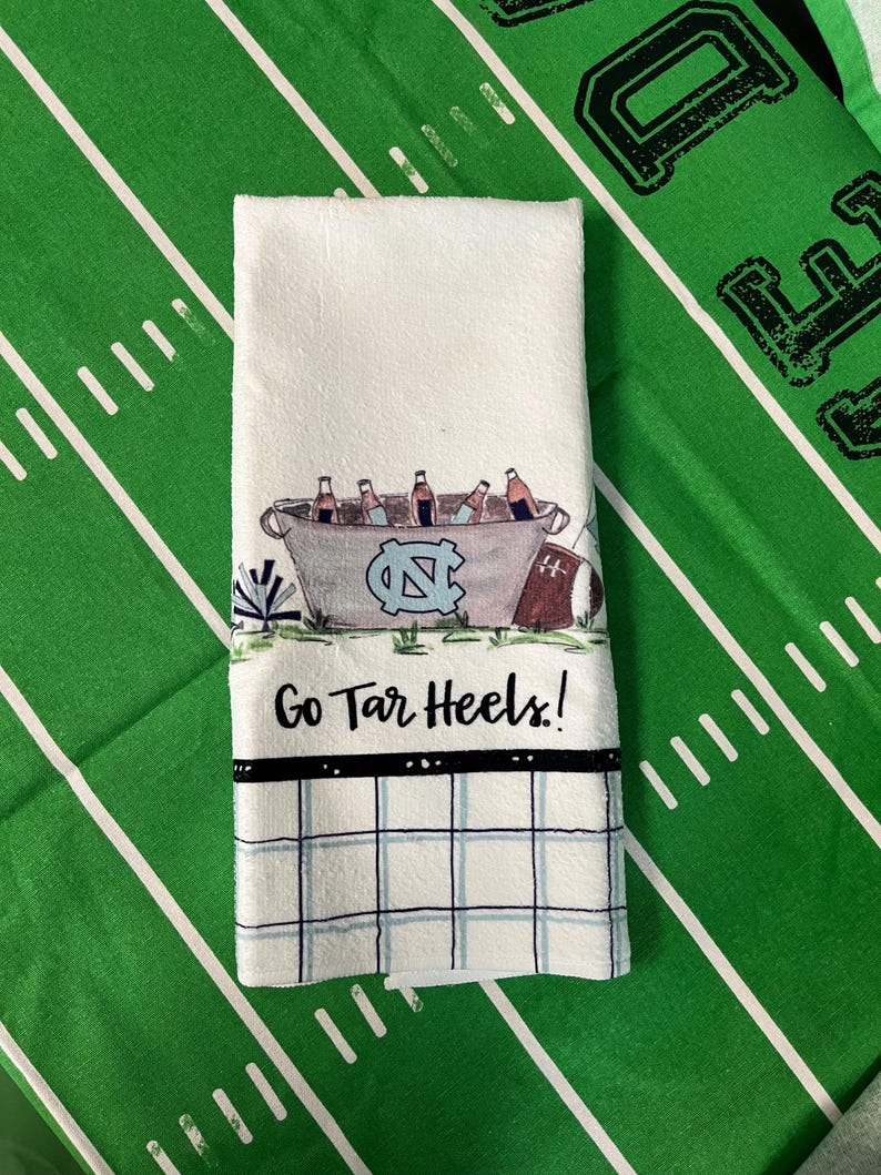 May include: White hand towel with a football field design. The towel features a bucket of beer bottles, the UNC logo, and the text "Go Tar Heels!". A black and blue checkered pattern is at the bottom.